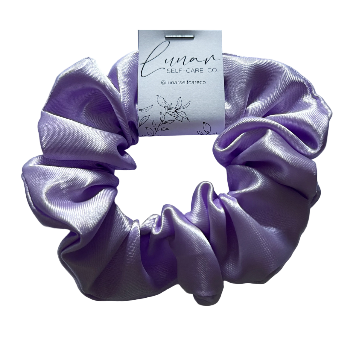 A lavender purple satin scrunchie with a white paper tag attached, displayed on a clean white background. The tag is branded 'Lunar Self-Care Co.