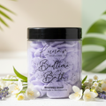 Jar of 'Bedtime Bath' whipped soap with lavender and flowers on a white surface with green leaves in the background