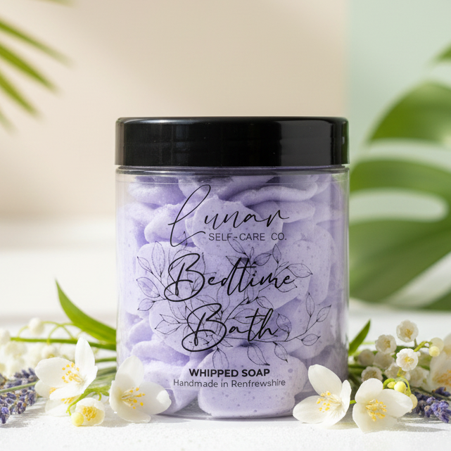 Jar of 'Bedtime Bath' whipped soap with lavender and flowers on a white surface with green leaves in the background
